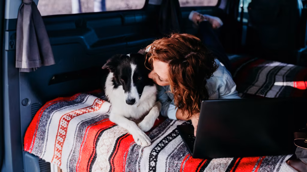 A woman relaxes with her dog and a laptop on a comfortable bed setup inside her vehicle to show that does sleeping in your car count as camping.