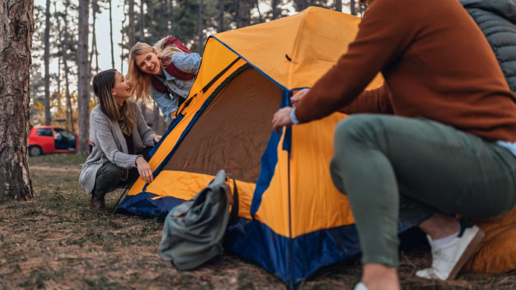 A group of friends laughing together while setting up a yellow tent shows how can camping be a source of building a good relationship through shared outdoor activities.