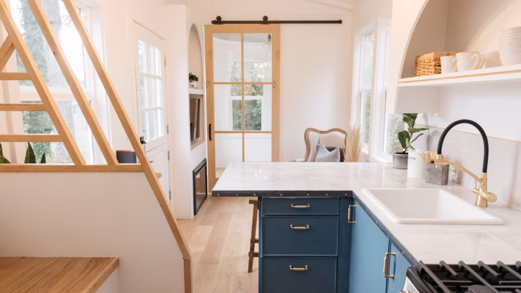 An interior view of a custom-designed tiny house on wheels showing features that address the question: can larger people live in a tiny house without sacrificing comfort. The home includes wide-plank light oak flooring, a generous L-shaped kitchen counter with a deep sink and full-sized range, and a wide interior walkway leading past a storage staircase. In the background, a large French door with clear panes and a sliding barn door provide ample light and a spacious, barrier-free access point. The high ceiling height and bright, open-concept tiny house floor plan contribute to a sense of physical comfort and ease of movement in a compact footprint.