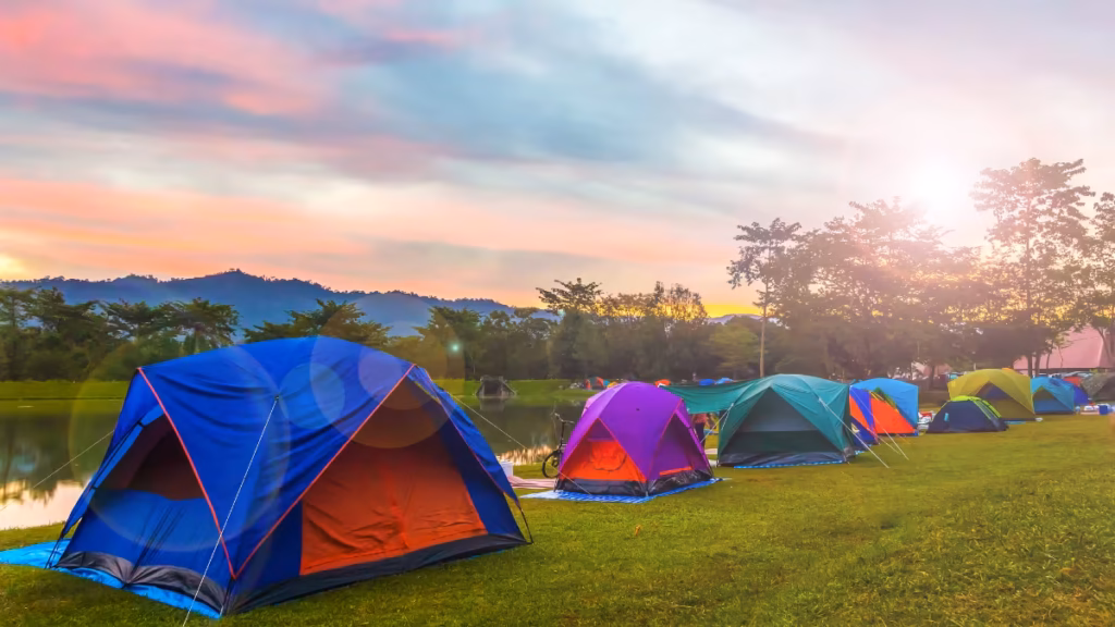 A row of colorful camping tents sits on a grassy field by a peaceful lake at sunset, illustrating how to tent camp without electricity in a scenic outdoor setting.