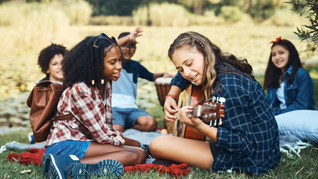 A group of girls laughing and playing guitar together at a lakefront campsite, showcasing the social bonding made possible by the fun things every girl should bring to summer camp.