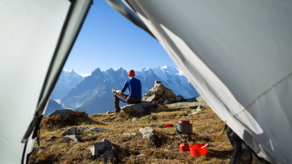 A solo traveler sits on a rocky ridge overlooking a mountain range to demonstrate why is it important to select a good campsite for both safety and views.