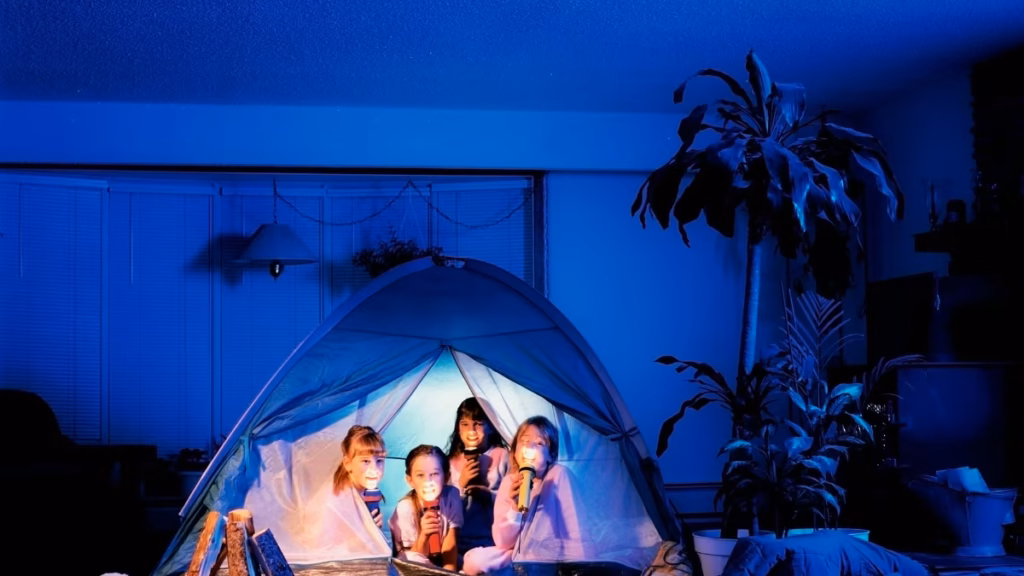 Four children holding flashlights inside a tent pitched indoors at night, illustrating DIY indoor camping ideas in a living room setting.