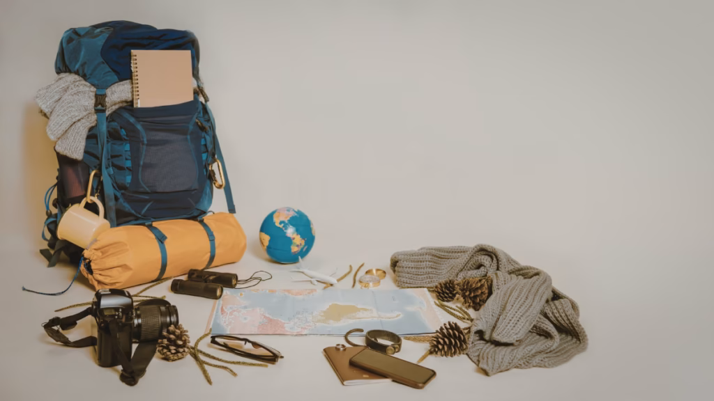 Camping backpack laid out with essential gear including sleeping mat, binoculars, map, and warm layers showing the key things to bring to summer camp.